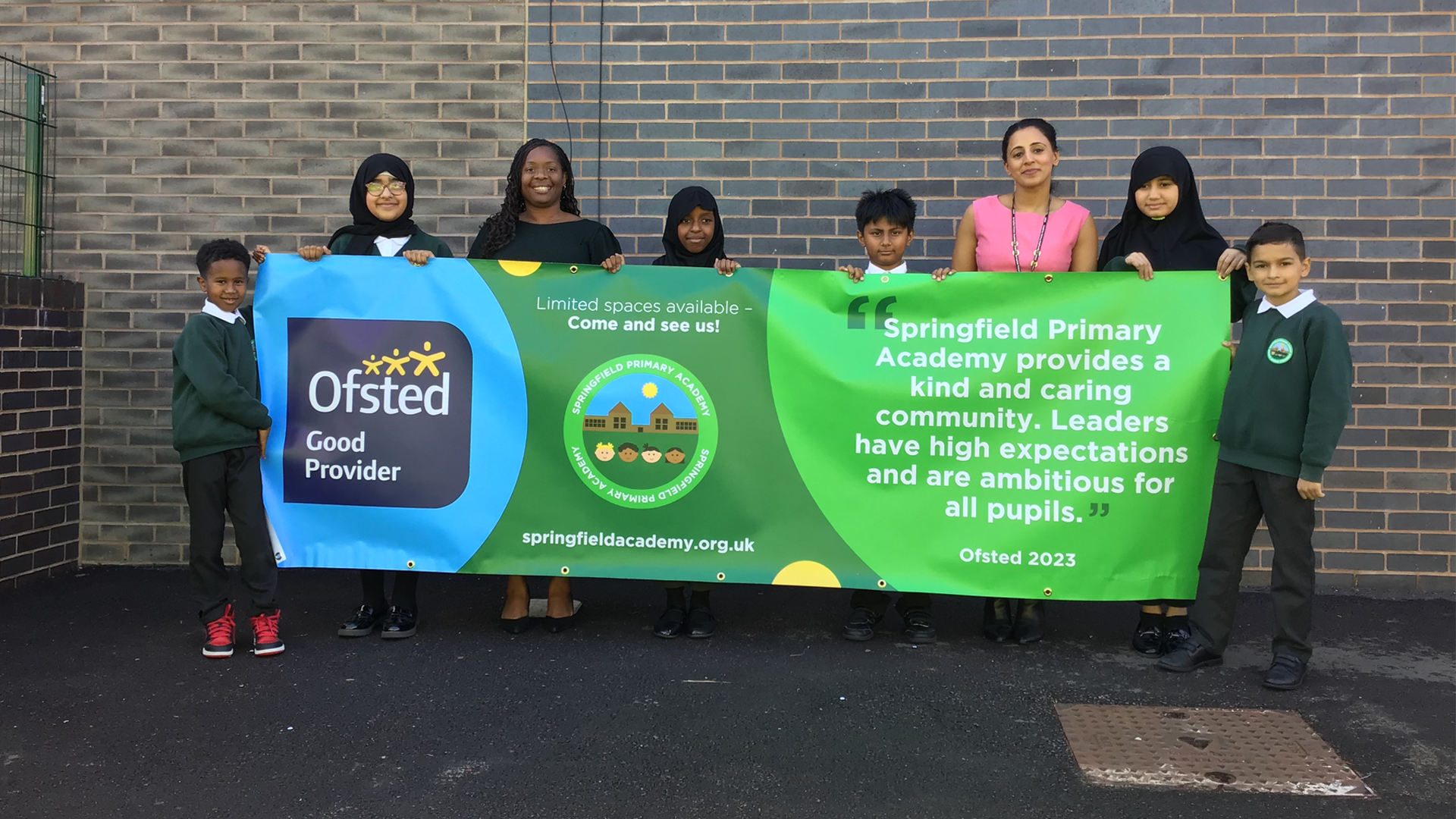 A photo of the children and staff at Springfield Academy holding a banner with a quote from the recent Ofsted Inspection which states "Springfield Primary Academy provides a kind and caring community. Leaders have high expectations and are ambitious for all pupils."